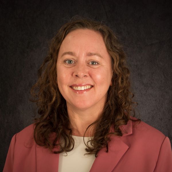 Smiling woman with curly hair in a pink blazer against a dark background.