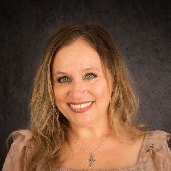 Smiling woman with blonde hair and a cross necklace against a dark background.