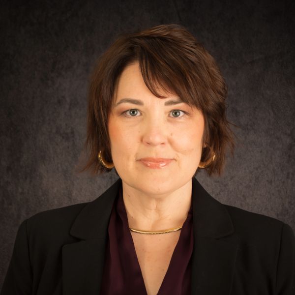 Professional woman with short brown hair in formal attire against dark background.