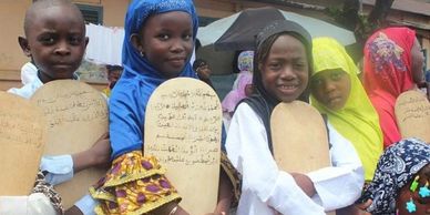Children holding wooden boards with Arabic script, dressed in colorful traditional attire.