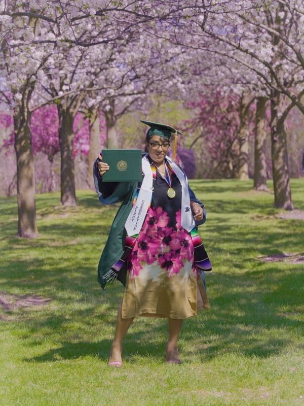 Graduate in cap and gown holding diploma under blooming cherry blossom trees.