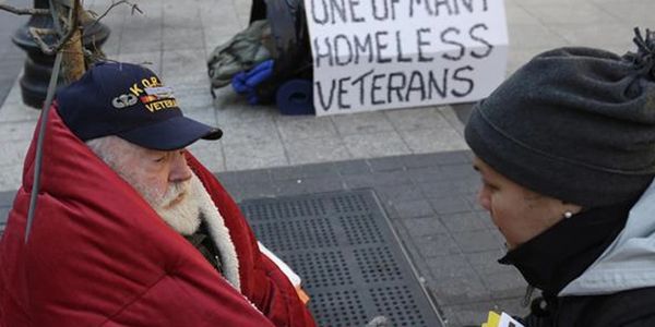 A homeless veteran wrapped in a red blanket speaks with a helper on the street.