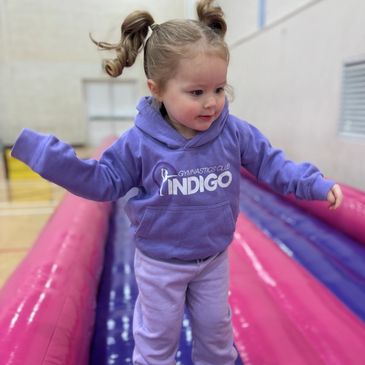 Toddler in purple hoodie playing on an inflatable gymnastics mat indoors.