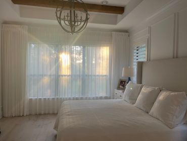 Sunlight fills a cozy, white bedroom with sheer curtains.