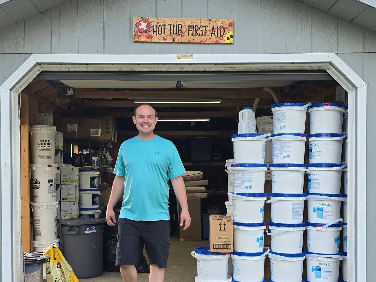 Man smiling in a storage shed with stacked white buckets and a "Hot Tub First Aid" sign above.