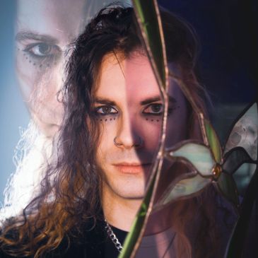 Person with long hair and dramatic makeup behind broken glass.