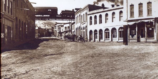 Historic town street with train on elevated track and horse-drawn carriage.