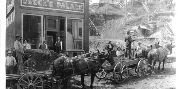 Old western town scene with horses, wagons, and people.