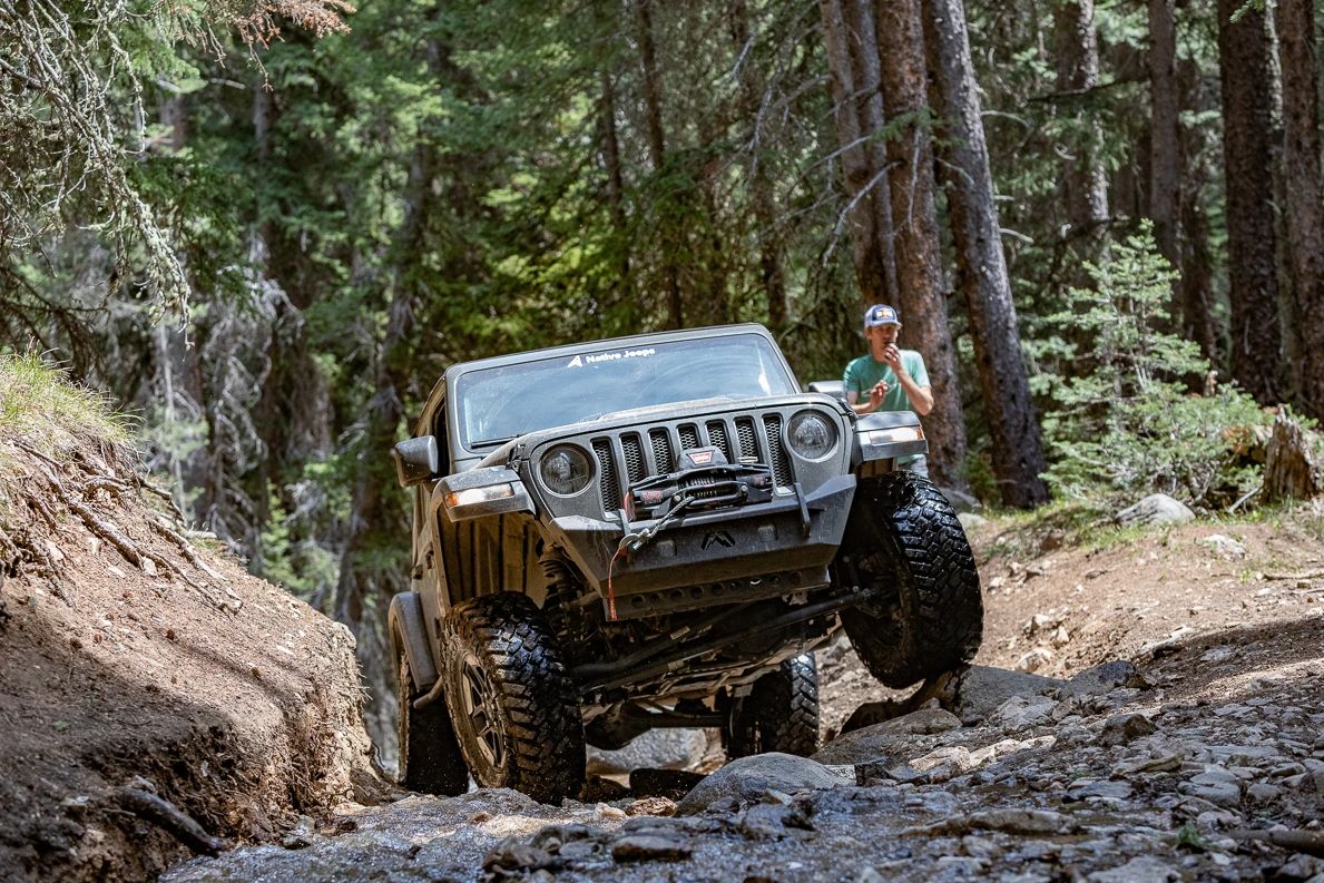 A Jeep maneuvering rocky terrain in a forest with a man observing.
