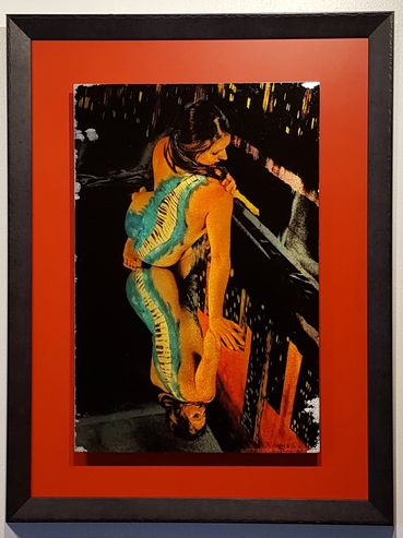 Woman facing away with blue piano painted down her back, seated on a mirror against a painted city