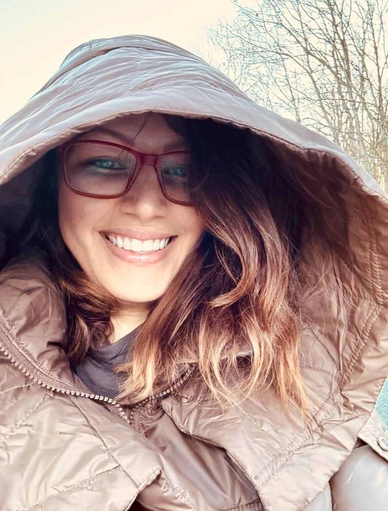 Smiling woman in a brown hooded jacket with glasses outdoors.