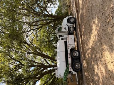 White tanker truck parked under large trees.