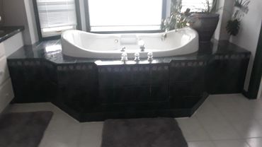 A white bathtub with black tile surround in a bathroom with rugs and plants.