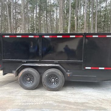 Black dual-axle trailer parked on concrete with forest background.