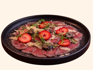 Beef carpaccio garnished with strawberries, herbs, and shaved cheese on a black plate.