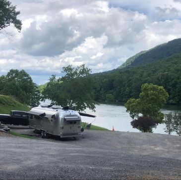 A silver camper trailer parked by a river with trees and hills in the background.