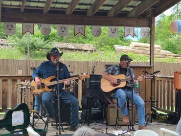 Two men playing guitars and singing on a wooden stage outdoors.