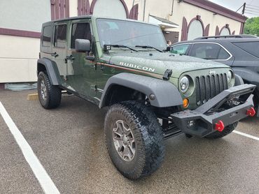 Green Jeep Rubicon with off-road tires parked in a wet lot.