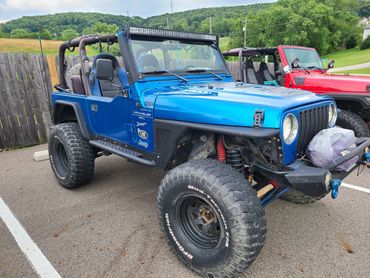 Blue Jeep Wrangler with off-road tires parked next to a red Jeep in a rural area.