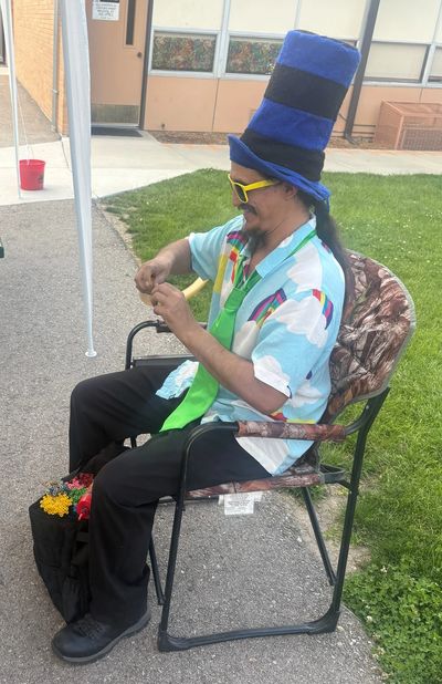 Man wearing a tall blue hat and colorful outfit sitting on a chair outdoors.