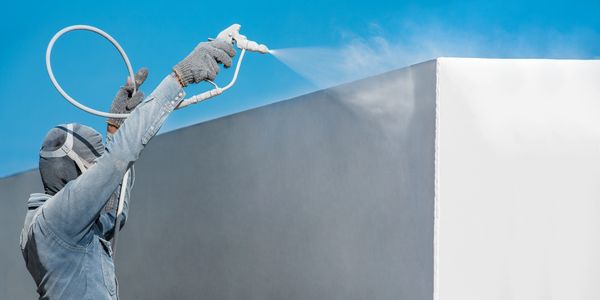 Worker spray painting a wall white with protective gear under clear blue sky.