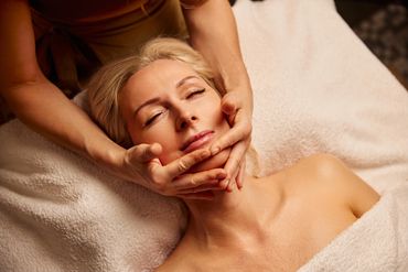 A person with closed eyes is lying on a white towel while receiving a rejuvenating facial massage.