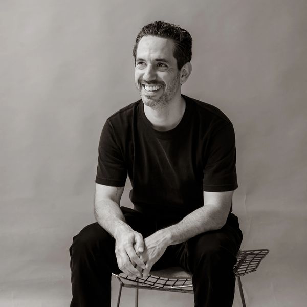 A man in black clothes sitting and smiling on a wire chair.