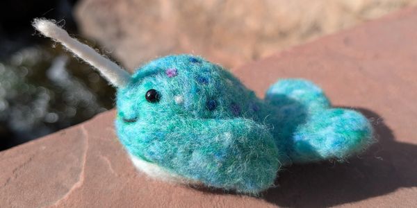 A small, handmade felt narwhal with blue and turquoise colors.