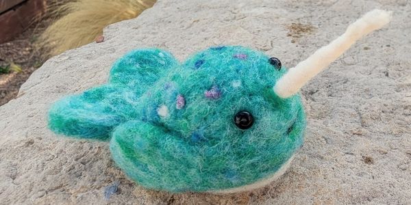 A handmade felted narwhal with blue and green colors and a white tusk.