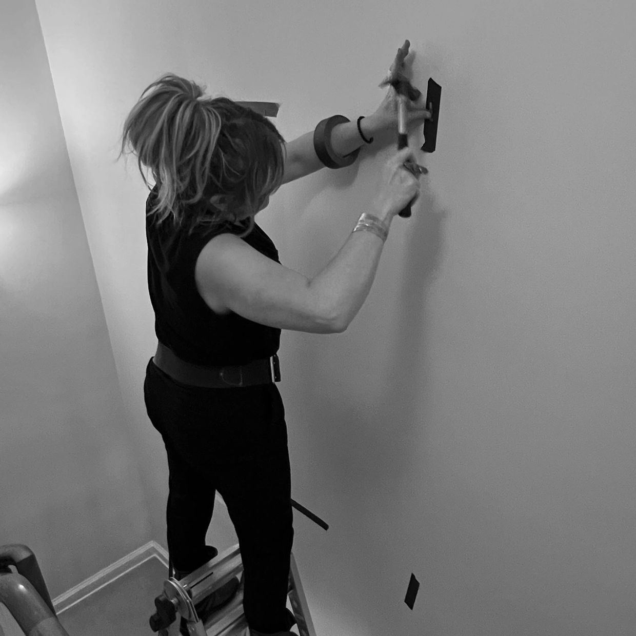 A woman standing on a ladder installing artwork mounting hardware.