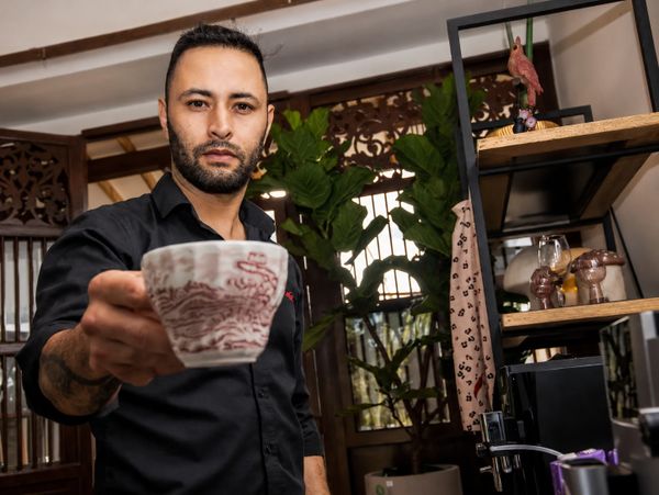 Man holding a patterned ceramic cup toward the camera indoors.