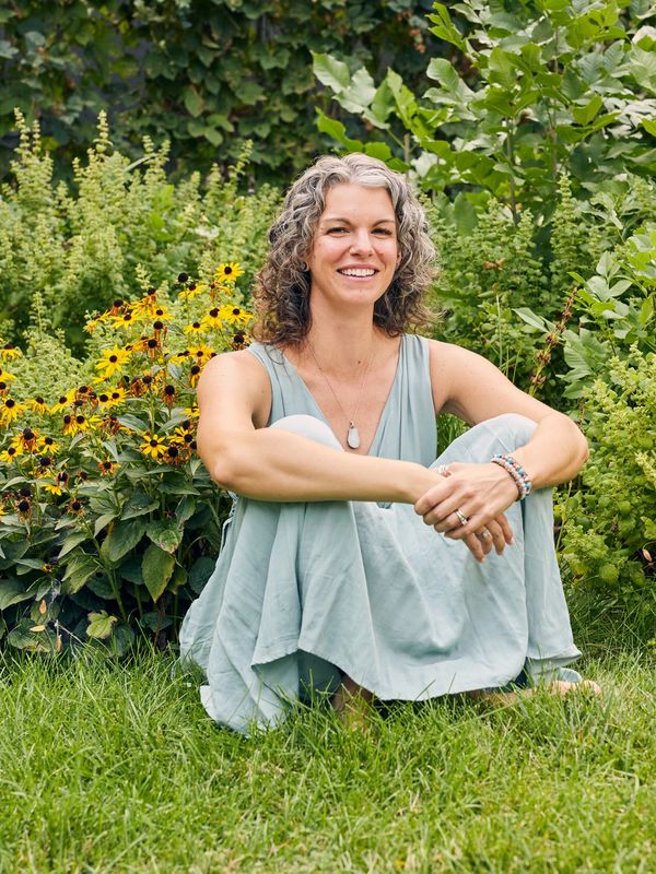 Woman with curly hair sitting on grass near yellow flowers, smiling.