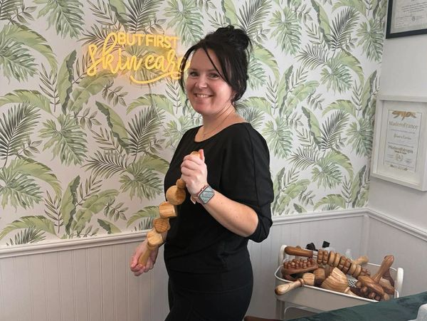 Smiling woman holding wooden massage tools in a spa room with tropical wallpaper.