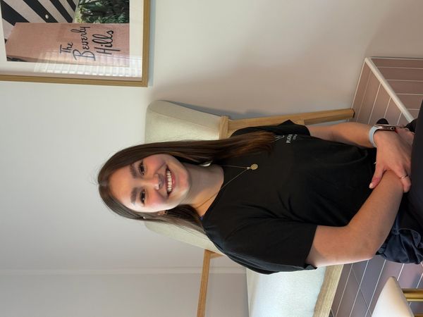 Smiling young woman sitting indoors with a framed Beverly Hills poster behind her.