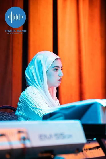 Woman in white hijab playing keyboard under vibrant stage lights.