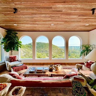 Sunlit living room with arched windows overlooking green hills and cozy seating.