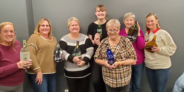 Seven women standing in a line holding various decorative bottles.