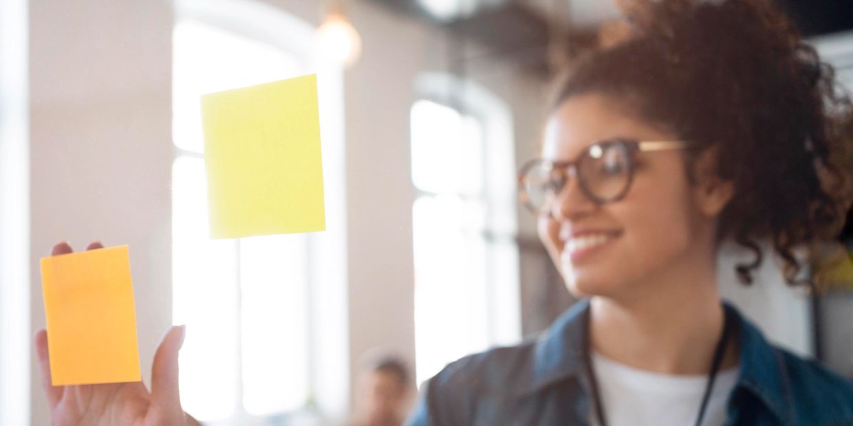 Mulher branca, veste blusa branca com um camisa azul, está colocando um post-it no vidro.