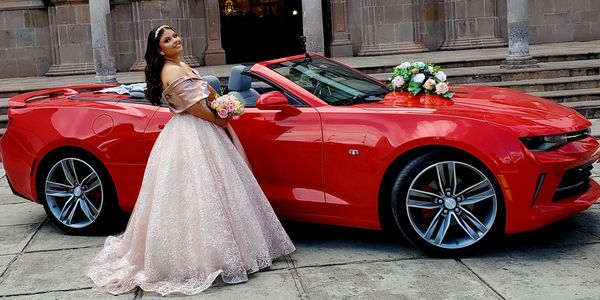 A woman in an elegant gown stands beside a red convertible decorated with flowers.