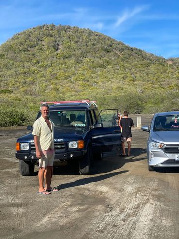 Man standing beside a blue Land Rover with a hill in the background.