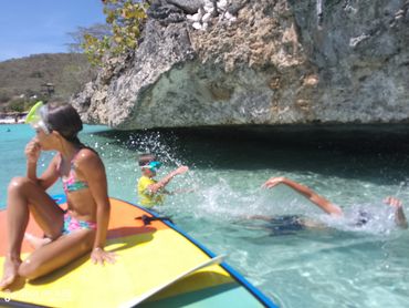 Children playing in clear water near a rocky shore, one on a paddleboard.