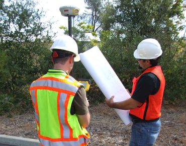 Two construction workers in safety gear examining blueprints outdoors.