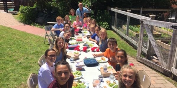 A group of children and an adult enjoying a meal outdoors at a long table.