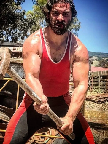 Muscular man in red tank top holding wooden stick.
