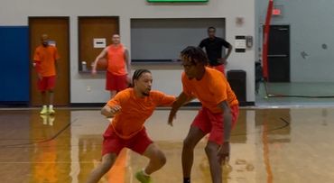 Two men playing basketball indoors, wearing orange shirts and red shorts.