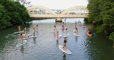 A large group of girls in bikinis paddle boarding in front of the Rainbow Bridge in Haleiwa
