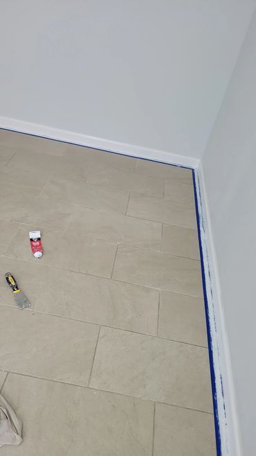 A corner of a room with tiled floor and a tube of adhesive and putty knife on the floor.