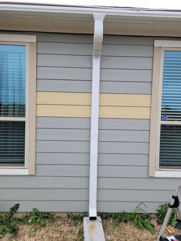 A white downspout on a gray and beige house exterior between two windows.