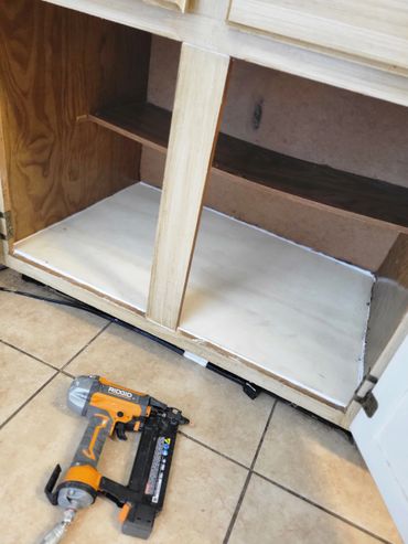 Empty wooden cabinet with a pneumatic nail gun on tiled floor.