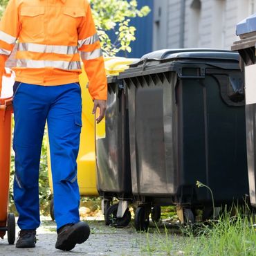 A valet trash worker walks and pulls a trash can behind him.